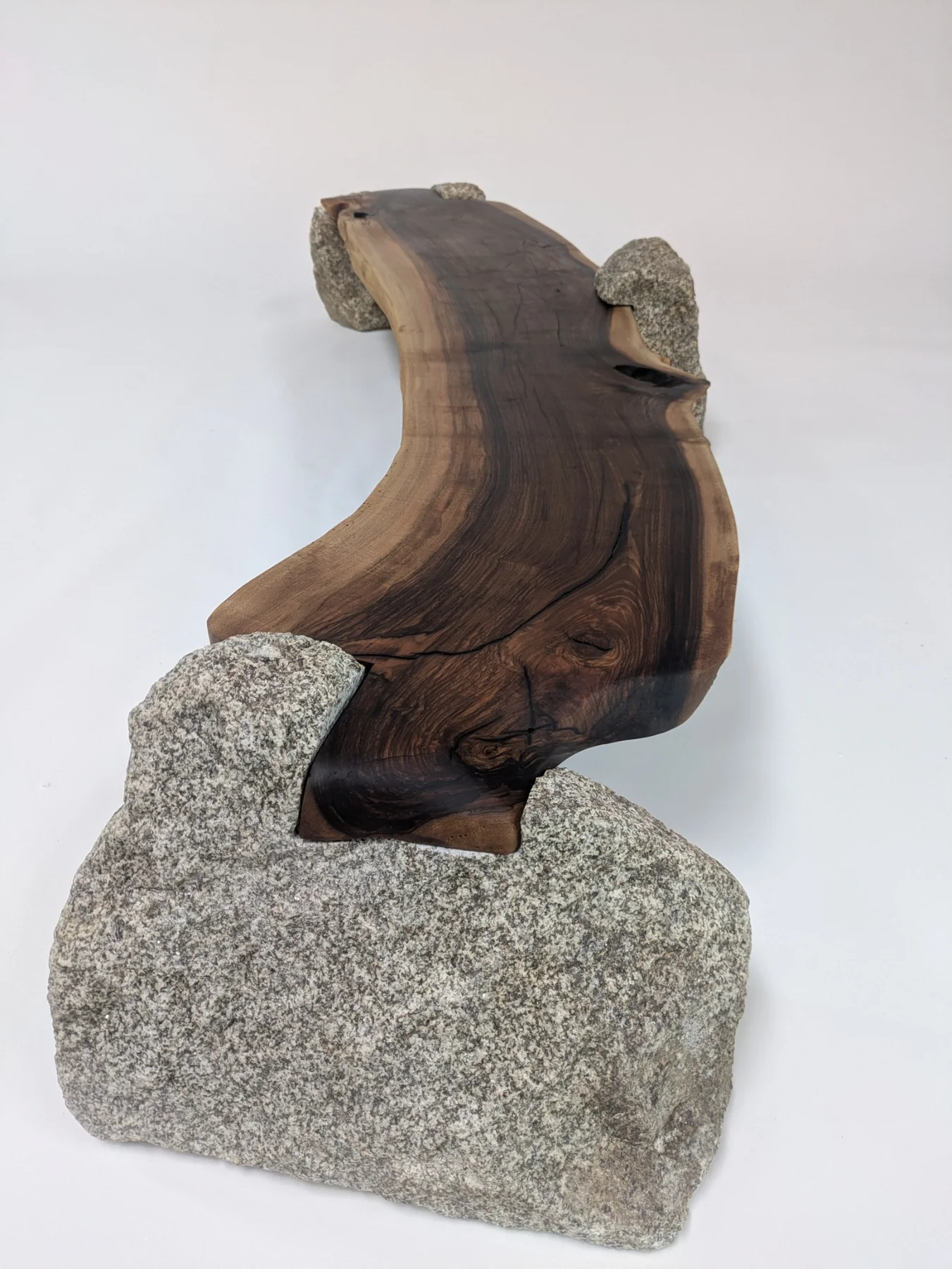 The curved walnut slab seen from one end between the granite stones