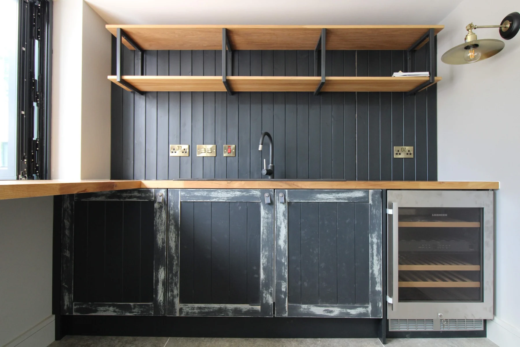 Distressed painted home bar doors beneath an oak worktop, with a panelled backing and metal-framed shelves