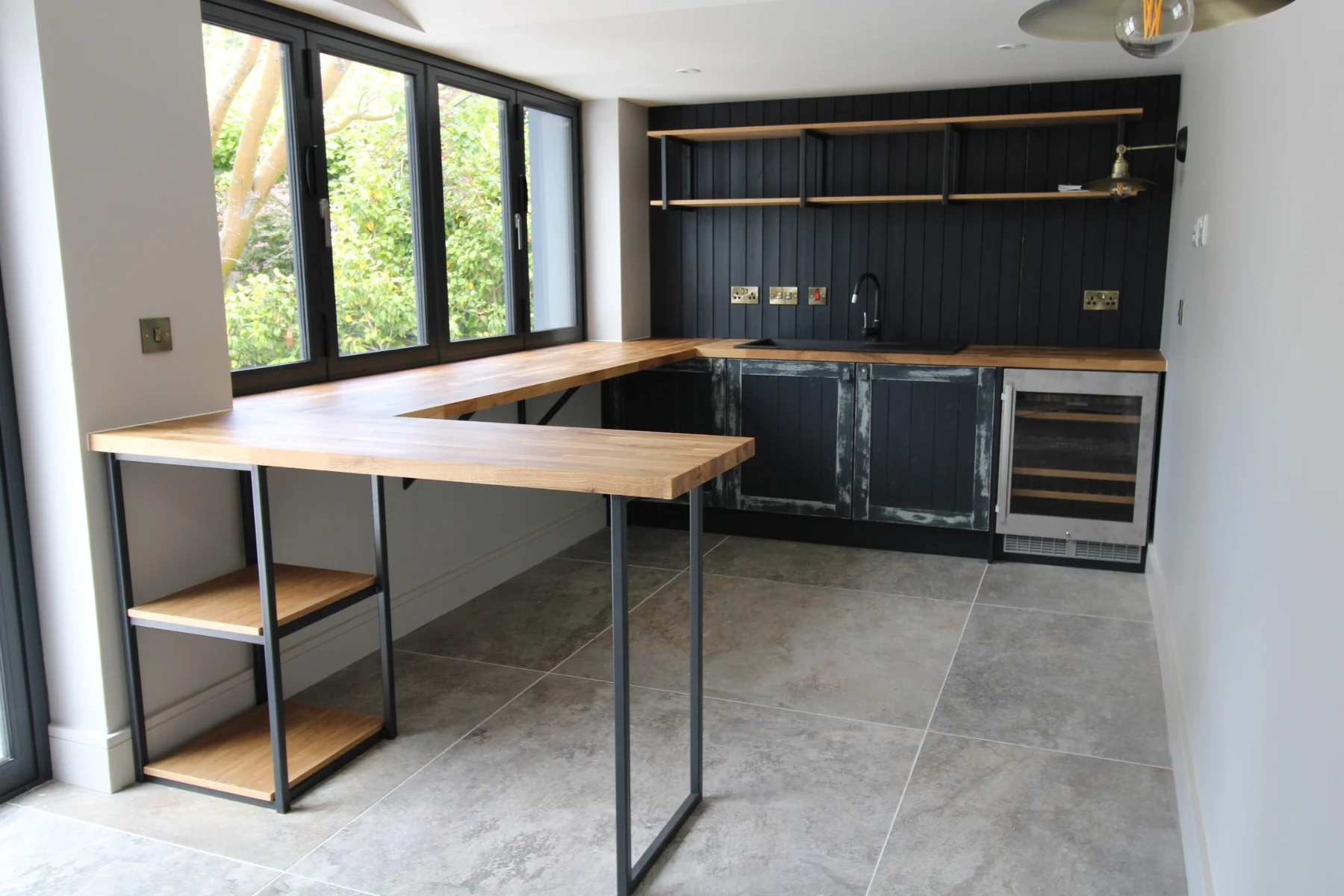 Home bar with a solid oak butcher-block worktop, black metal supports and distressed painted cupboard doors